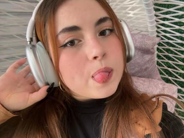 Close-up of VicluX, a young woman with light skin and brown hair, wearing silver headphones, sticking out her tongue. She has dark eyeliner and is wearing a black top and tan coat.