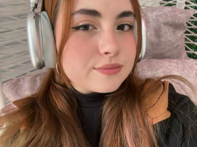 Close-up of VicluX, a fair-skinned woman with long brown hair, wearing large silver headphones and a black turtleneck, leaning back with a relaxed expression, light pink pillow behind her.