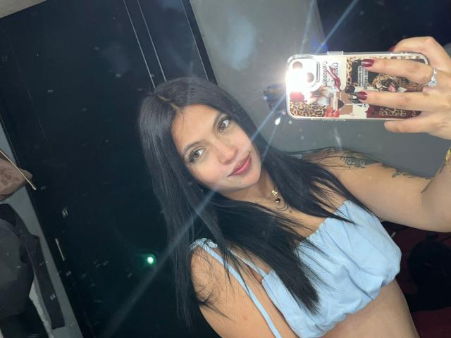 NyraVanel, a hot woman with long black hair, takes a selfie in a mirror. She's wearing a white crop top, holding a glittery phone, smiling seductively. Bright light flares.