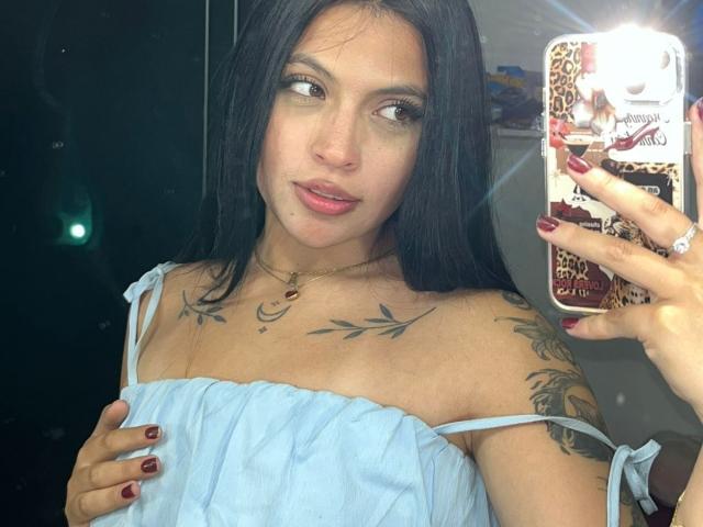 NyraVanel, with long black hair, gazes at her phone, wearing a white off-shoulder top, showing tattoos on her chest and arm, holding her phone with red nails, reflecting light.