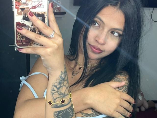 NyraVanel se toma un selfie con su mano derecha, mostrando un tatuaje floral negro detallado en su antebrazo. Ella lleva una camiseta blanca con tirantes, uñas rojas y una pulsera dorada. Muy atractivo..