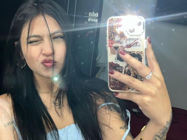 NyraVanel, une jeune femme aux longs cheveux noirs, prend un autoportrait avec le flash. Elle porte un débardeur blanc, a du vernis à ongles rouge et une bague à son doigt. Extrêmement sexy..