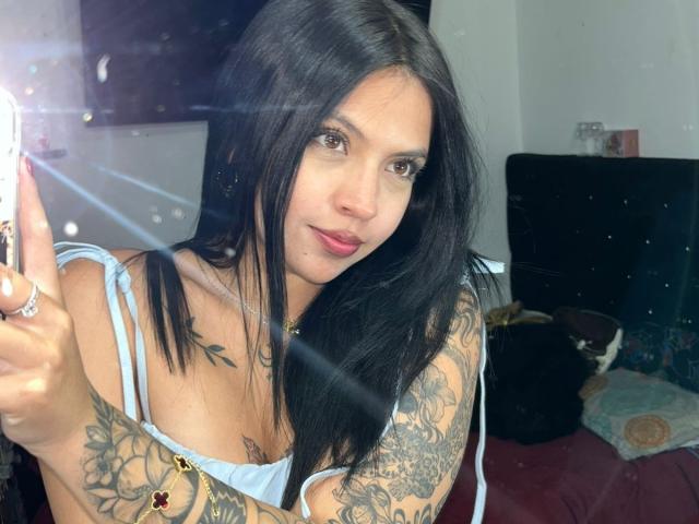 Gros plan de NyraVanel, une femme séduisante avec de longs cheveux noirs, des tatouages sur les bras et un soutien-gorge blanc. Elle fait un signe de paix, un éclair de lumière, avec un sourire séduisant..