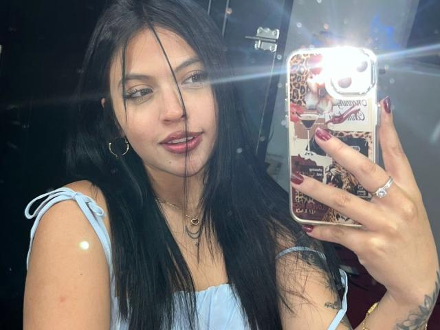 NyraVanel, une jeune femme aux longs cheveux noirs, prend un autoportrait avec un smartphone lumineux. Elle porte un haut bleu clair, des boucles d'oreilles pendantes, et a des ongles rouge foncé, tout en affichant un sourire discret..