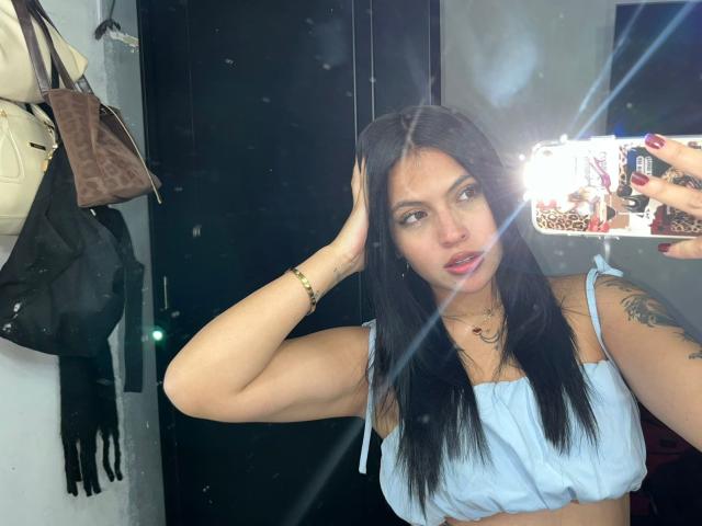 NyraVanel, avec ses longs cheveux noirs, porte un crop top blanc et se tient la tête d'une main, tout en prenant un selfie. Un éclair lumineux se reflète sur son visage, des sacs sont suspendus en arrière-plan..