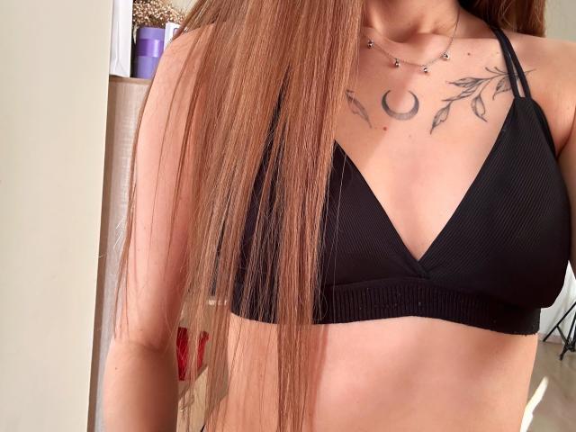 Primer plano de Karish con cabello castaño largo y liso, vistiendo un top corto negro con textura, que revela una luna creciente y una hoja. Tatuaje en su pecho. Sus pezones son visibles a través de la tela..