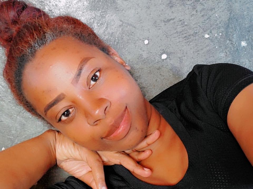 Close-up photo of Akanny69 with brown skin, red hair in a bun, and a black shirt. She's leaning on her hand, smiling subtly, with visible facial acne and a relaxed pose.