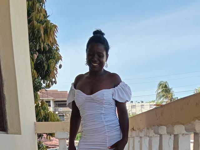 Photograph of a dark-skinned woman with a curvy physique, wearing a white off-shoulder dress, standing on a balcony. She has her hair in a bun, smiling. Ocean in the background. Fucking hot.