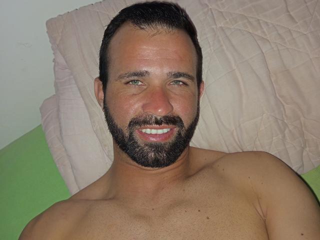 Hector Sales, sin camisa, con barba completa y cabello negro corto, acostado sobre un almohadón blanco, sonriendo con un ligero rubor. piel, vello visible en el pecho y un fondo de sábana verde. Muy atractivo..