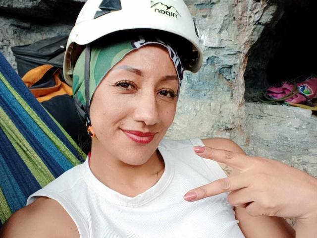 Foto van SamanthaCorine met een gebruinde huid, die lacht, gekleed in een wit tanktop en een witte helm, een groene hoofddoek, en met een vinger. wijzend naar de camera, leunend tegen een muur met een structuur, kleurrijke gestreepte stof op de achtergrond..