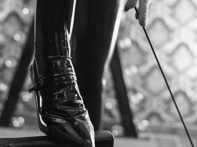 Fotografía en blanco y negro de las brillantes botas de charol de la marca Mistress-Joy-hot, que le llegan hasta la rodilla y tienen cordones, mientras está de pie en un escenario. Sosteniendo un látigo de equitación en una mano. Luces de fondo desenfocadas con efecto bokeh..
