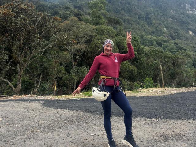 SamanthaCorine está em um caminho de cascalho, vestindo um moletom vermelho, jeans azuis e um capacete branco. Ela está levantando a mão direita, com uma parede de pedras ao fundo. Pose incrivelmente sexy..