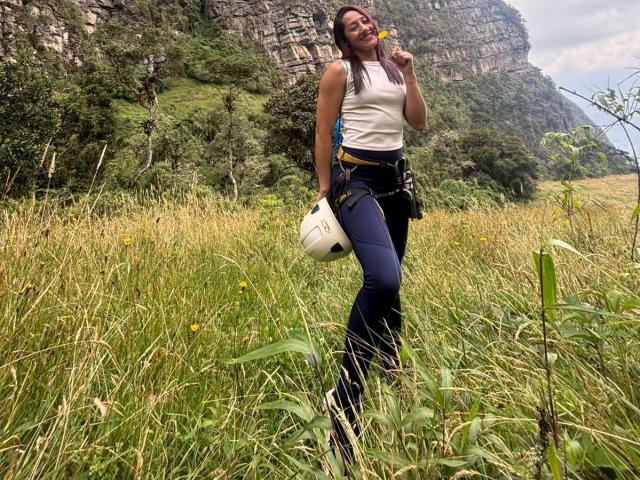 SamanthaCorine steht auf einem grasbewachsen Feld, hält eine weiße Tasche in einer Hand und eine gelbe Blume an ihren Mund. Sie trägt ein weißes Tanktop und blaue Jeans, im Hintergrund sind eine felsige Klippe und Bäume zu sehen. Sie ist unglaublich schön..