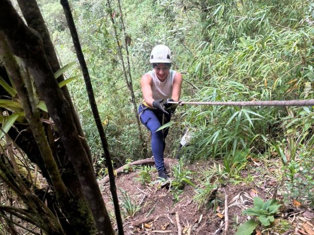 SamanthaCorine se agacha en una jungla densa y verde, vistiendo un casco blanco y una camiseta sin mangas, pantalones azules y zapatos. Está concentrada, con una cuerda frente a ella, creando tensión..