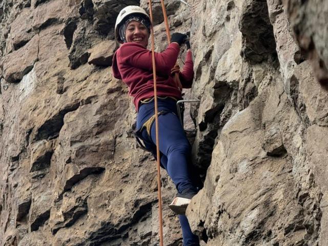 SamanthaCorine, che indossa un casco bianco e una maglietta rossa, scala una parete rocciosa con una corda. Pantaloni blu aderenti, scarpe da arrampicata. Stringe la corda, sorridendo, mostrando tensione sessuale e determinazione..