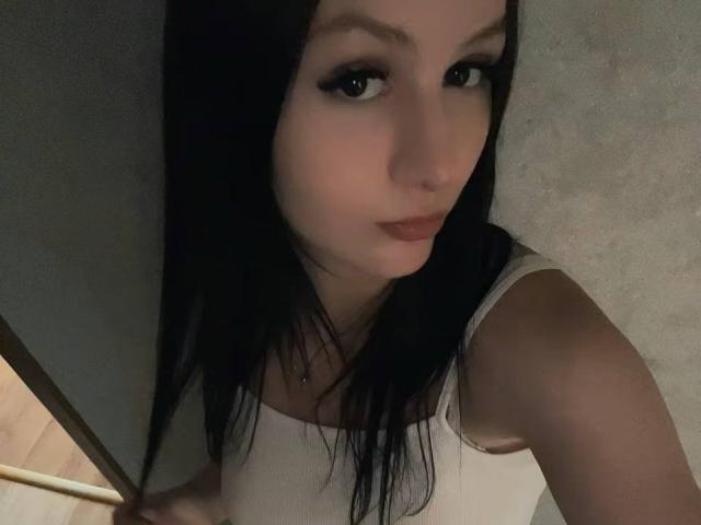 Close-up of LaylaArchibeque, a woman with pale skin and black hair, wearing a white tank top, looking seductively at the camera, holding a golden object. Dimly lit, sexual tension.