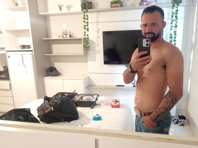 Hector, torse nu, avec une barbe soignée et des tatouages sur son bras droit, tenant un téléphone, debout dans une pièce lumineuse. jeans ouverts, laissant entrevoir un aperçu de son sexe, sac noir en désordre sur un bureau blanc..