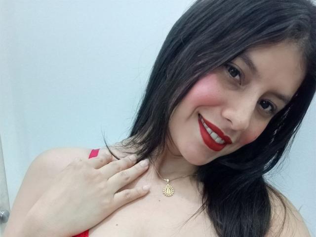 MadissonFontaine with dark, straight hair, red lipstick, and red top, smiling at the camera. Hand on chest, wearing a gold necklace. Soft light, slight angle, white background. Fucking sexy.