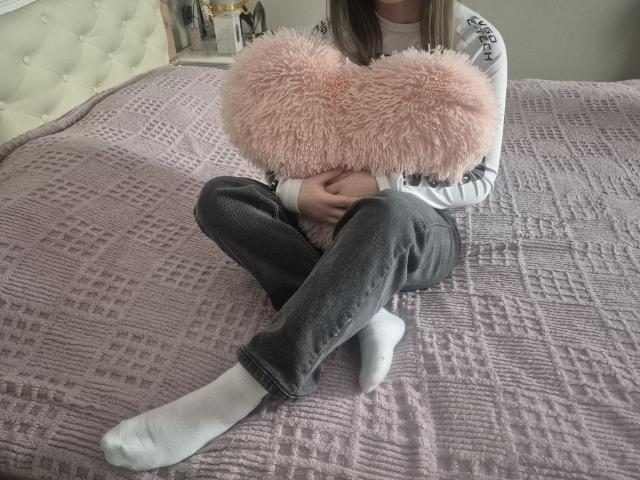 ZoieHurt sits on a purple textured bedspread, legs crossed, holding a pink fluffy heart pillow over her chest. Wearing black pants, white socks, and a white long-sleeve shirt.