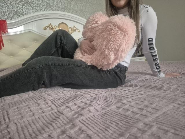 ZoieHurt è seduta su un letto con una coperta grigia e strutturata, tiene un cuscino rosa e soffice, indossa una maglia bianca a maniche lunghe e jeans neri, con le gambe divaricate, creando tensione sessuale.