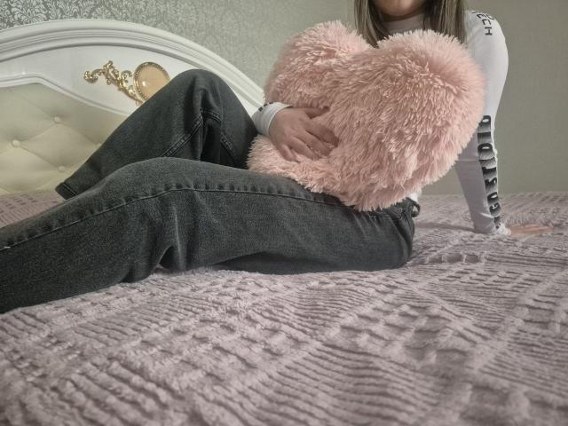 ZoieHurt, liggend op een bed, gekleed in zwarte jeans en een wit shirt, met een roze, pluizige hartvormige kussen in haar handen, benen gespreid. apart, het creëren van seksuele spanning, grijze, gestructureerde deken, wit, versierd hoofdeinde.