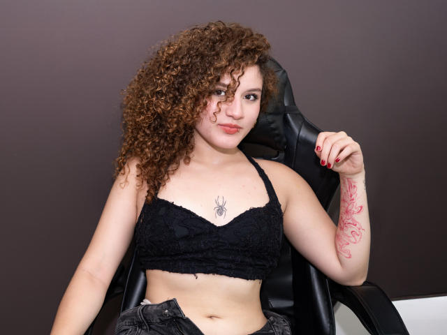 Fotografía de SelenaNova, sentada en una silla negra, vistiendo un sujetador de encaje negro, con cabello castaño rizado y uñas rojas. pulido y un tatuaje rojo en su antebrazo. Sonriendo, con la mano en la silla.