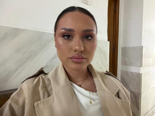 Photograph of AvalonWest with dark hair pulled back, wearing a beige jacket over a white top, gazing directly at the camera. Her skin is shiny, with pronounced makeup. Sexual tension evident.