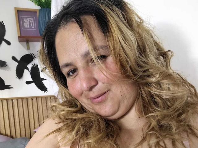 Primer plano de RouseWright, una mujer robusta con cabello castaño ondulado y con reflejos rubios, sonriendo ligeramente y mirando directamente a la cámara. Sus hombros desnudos son visibles, con calcomanías de pájaros negros en el fondo..