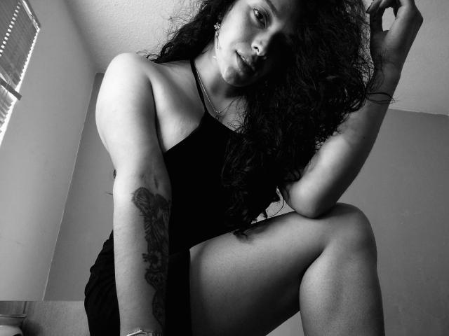 Fotografia in bianco e nero di Chantal Bernadette con i capelli ricci, che indossa un abito nero, seduta con le gambe incrociate, con una mano. capelli, tatuaggio sull'avambraccio, sguardo intenso, alto contrasto, tensione sessuale.