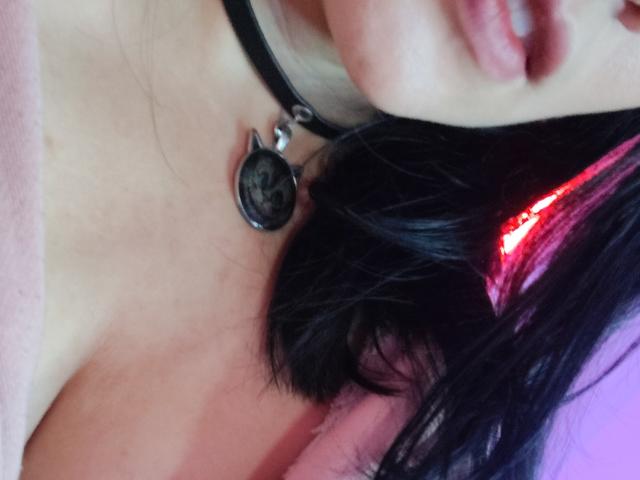 Primer plano del rostro y cuello de IssisDiosa, mostrando cabello negro con reflejos rojos, un collar negro con un colgante circular. y piel pálida. Centrado en la clavícula, labios ligeramente entreabiertos..