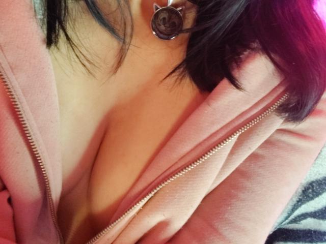 Close-up of IssisDiosa's chest and neck, wearing a pink zip-up hoodie with zipper partially open, exposing cleavage. Dark hair, silver hoop earring visible. Background blurred, focusing on skin and fabric.