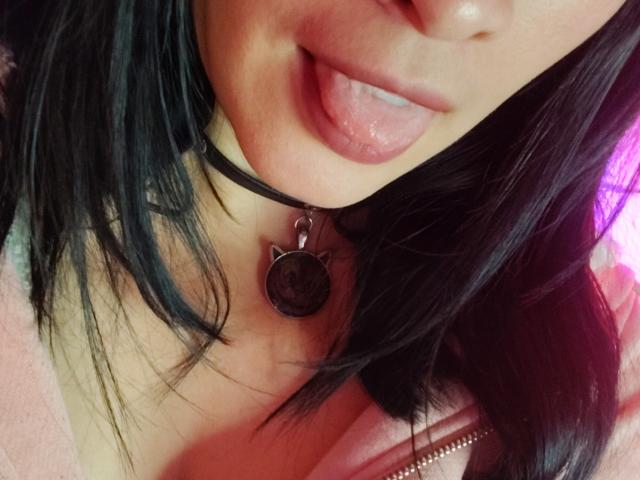 Primer plano del rostro y cuello de IssisDiosa, mostrando cabello negro, labios rosados con la lengua ligeramente fuera, un collar negro con un dije redondo. collar y chaqueta rosa. Ligera tensión sexual, ambiente provocador..