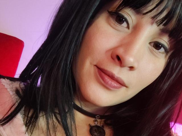 Close-up photo of IssisDiosa, a woman with straight black hair, bangs, and light skin, wearing a black choker with a pendant, gazing directly at the camera with a subtle smile. Background includes a red object.