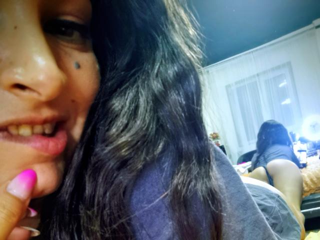 Close-up de PassionX com cabelo preto ondulado, pele morena e uma pinta na bochecha, sorrindo e mostrando um dedo com unhas pintadas de rosa. tocando seus lábios. O fundo mostra outra pessoa com cabelo escuro..