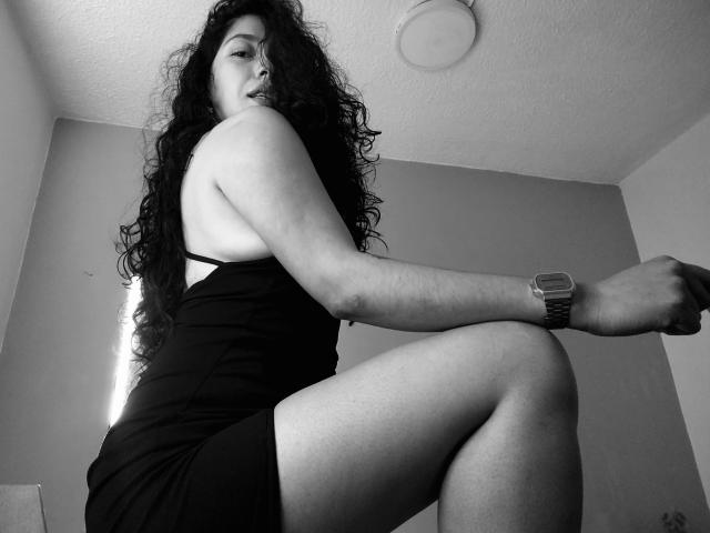 Black and white photo of ChantalBernadette, with long curly hair, wearing a tight black dress, sitting with one leg raised, exposing her thigh, wearing a wristwatch, shadowed face, sexual tension.