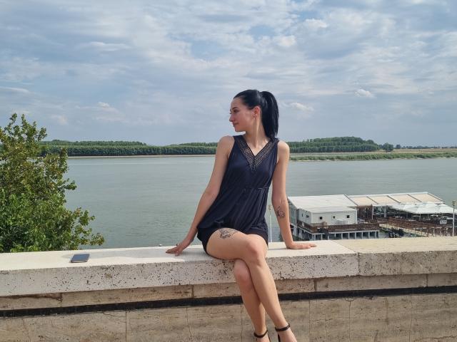 RoxxanneBoby, avec une queue de cheval foncée, une robe noire sans manches et des talons noirs, assise sur une corniche en pierre, les jambes croisées, avec un tatouage. Sur la cuisse droite, regardant vers la gauche, un lac et des bateaux en arrière-plan..