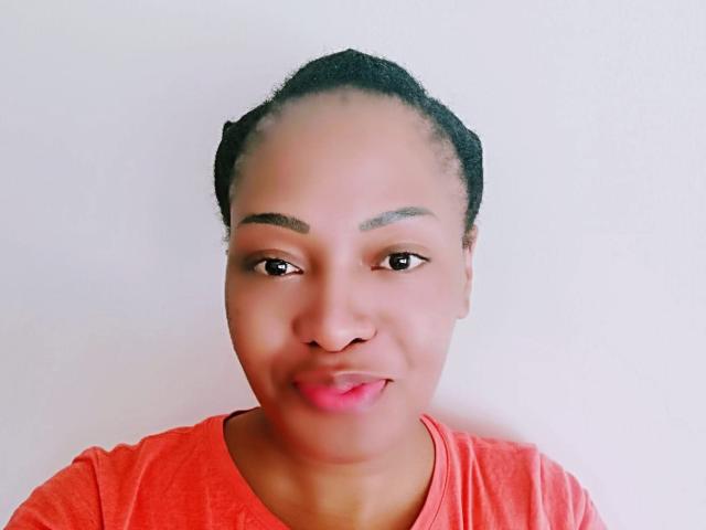 Photograph of a black woman with medium brown skin, dark hair pulled back, wearing a bright orange shirt. She smiles slightly, looking directly at the camera, with a white background. Her eyebrows are thick and arched. She has a subtle, confident expression, with a hint of sexual tension.