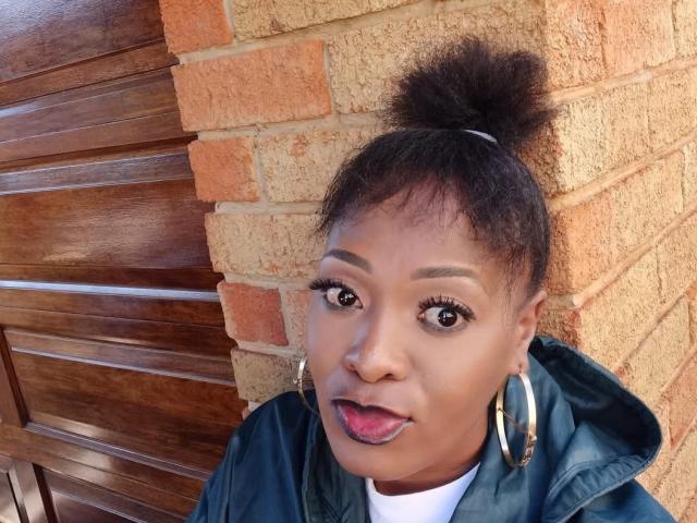 SparkleJoy, black woman with dark skin, big eyes, and thick eyebrows, tongue out, black hair in a high bun, wearing large hoop earrings, green jacket, brick wall, wooden door. Fucking hot.