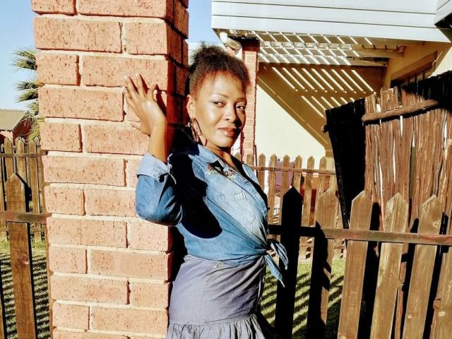 SparkleJoy, a Black woman with short curly hair, leans against a brick wall, waving with a flirtatious smile. She wears a blue button-up shirt tied at the waist, standing by a wooden fence. Bright sunlight.