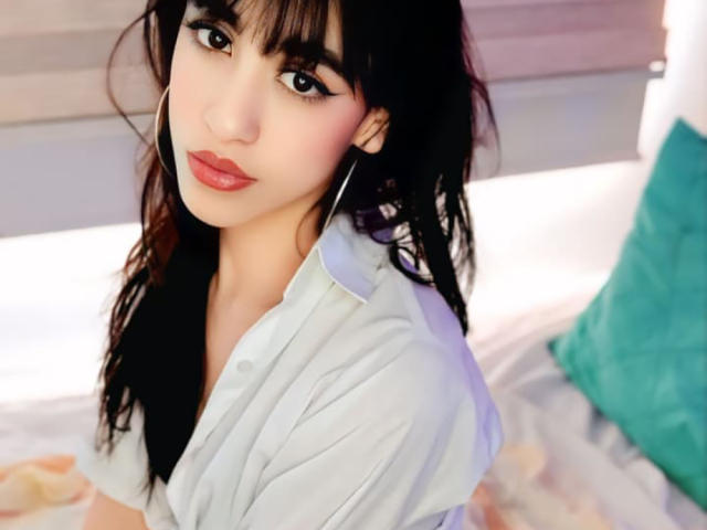 Fotografia ravvicinata di VanellopeLm, una donna attraente con lunghi capelli neri, rossetto rosso e occhi scuri, che indossa una camicia bianca. seduta su un letto con un cuscino turchese, che guarda seducentemente verso la fotocamera.