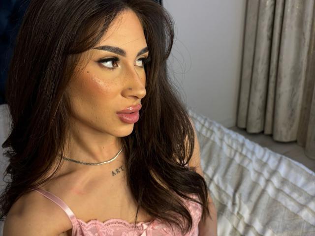 Fotografía de primer plano de SofiyaXO, una mujer de cabello oscuro con una piel radiante, labios rosados y un delineador de ojos marcado. Ella mira de lado con una expresión seductora, vistiendo una blusa rosa de encaje. La luz entra desde la derecha.