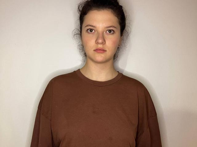 AshleyMonas heeft bleke huid en donker haar in een rommelige knot. Ze draagt een bruine sweater en staat voor een effen witte achtergrond, recht vooruit kijkend met een neutrale uitdrukking. uitdrukking.