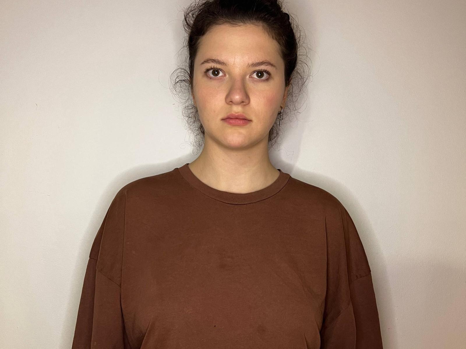 AshleyMonas ha la pelle chiara e i capelli scuri raccolti in un'acconciatura disordinata. Lei indossa una felpa marrone, è in piedi di fronte a uno sfondo bianco uniforme e guarda dritto davanti a sé con un'espressione neutra. espressione.