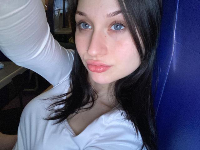 Selfie de perto de RoxxanneBoby, de pele clara, com olhos azuis e cabelo preto, vestindo uma blusa branca justa, com um leve decote. braço levantado, olhar direto, lábios rosados, tensão sexual, luz natural intensa..
