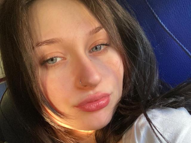 Primo piano di RoxxanneBoby, una donna dalla pelle chiara, con lunghi capelli neri, occhi azzurri e un piercing al naso. Sta guardando direttamente l'obiettivo con un sorriso leggero e seducente. La luce naturale e intensa mette in risalto il suo viso..