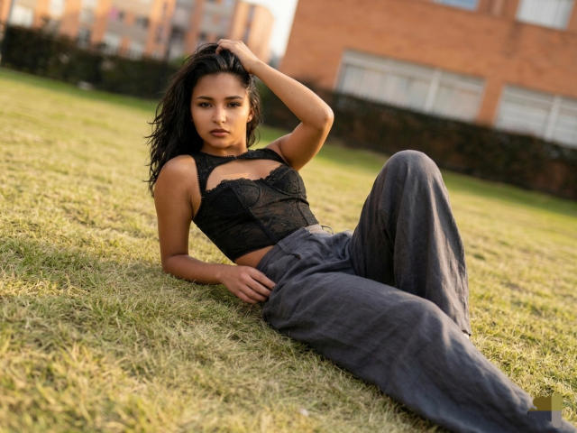 BellaChaudAmour est allongée sur l'herbe, les jambes écartées, portant un haut en dentelle noire et un pantalon gris ample. Elle regarde l'appareil photo, une main dans les cheveux, créant une tension sexuelle. Fond éclairé par le soleil.