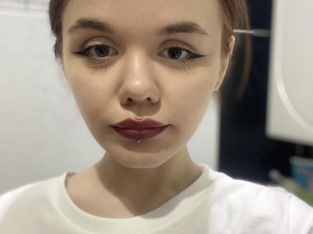 ShannaStorrer's face with red lipstick, dark winged eyeliner, and a small drop of saliva on her lower lip, wearing a white shirt, brown hair tied back.