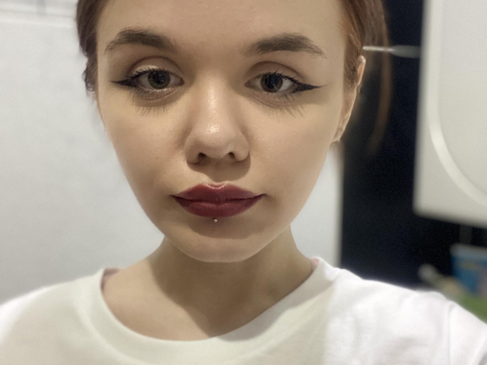 ShannaStorrer's face with red lipstick, dark winged eyeliner, and a small drop of saliva on her lower lip, wearing a white shirt, brown hair tied back.