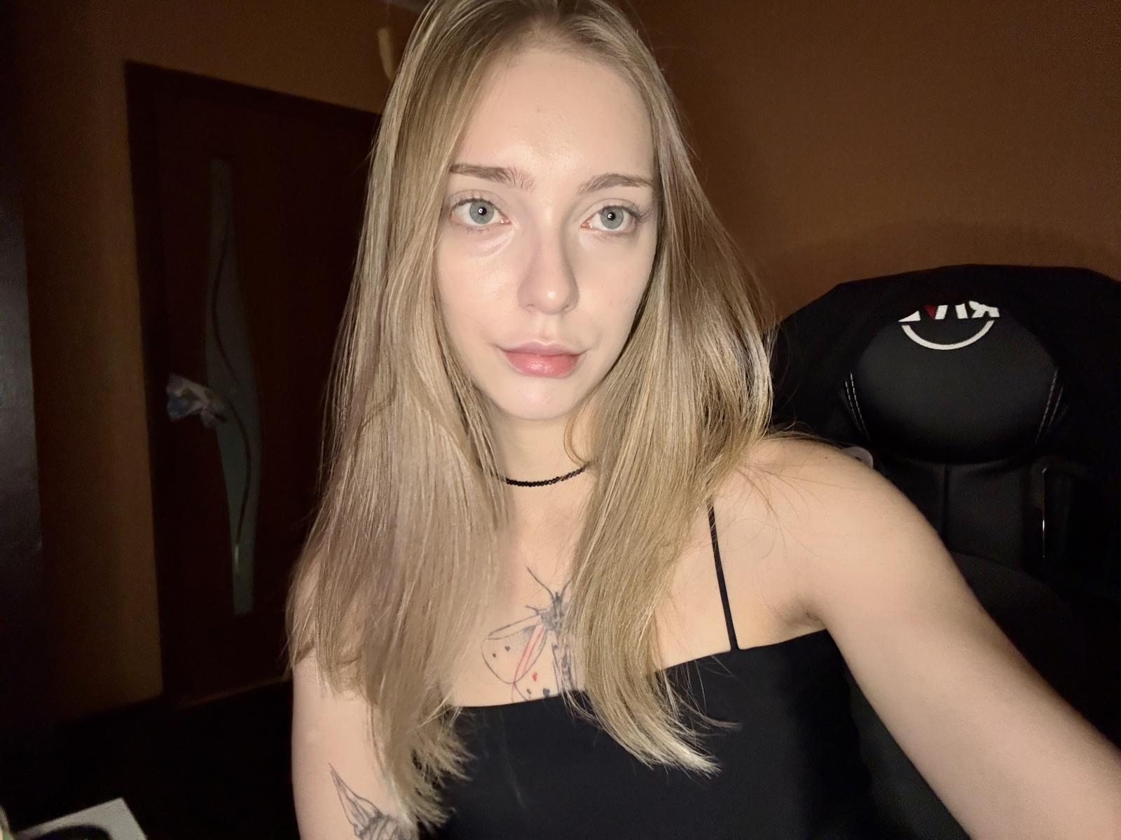 EdrisLueder has straight, light brown hair, pale skin, and blue eyes. She's wearing a black spaghetti strap top, exposing a colorful chest tattoo. Bright lighting highlights her face, creating intense focus.