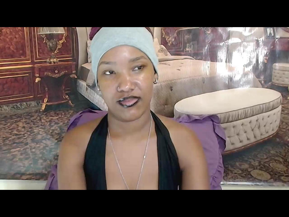 Fotografia di EroticSexyPrada con la pelle color marrone chiaro, che indossa un foulard bianco, un top nero a halter, una collana d'argento e orecchino, posizionato di fronte a un divano beige circolare e imbottito e a un mobiletto in legno scuro..